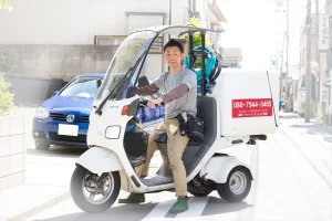 代表の田中次郎が清掃道具を積んだ三輪バイクにまたがり、電話番号とエアコン・ハウスクリーニングの文字が書かれた箱を載せている様子