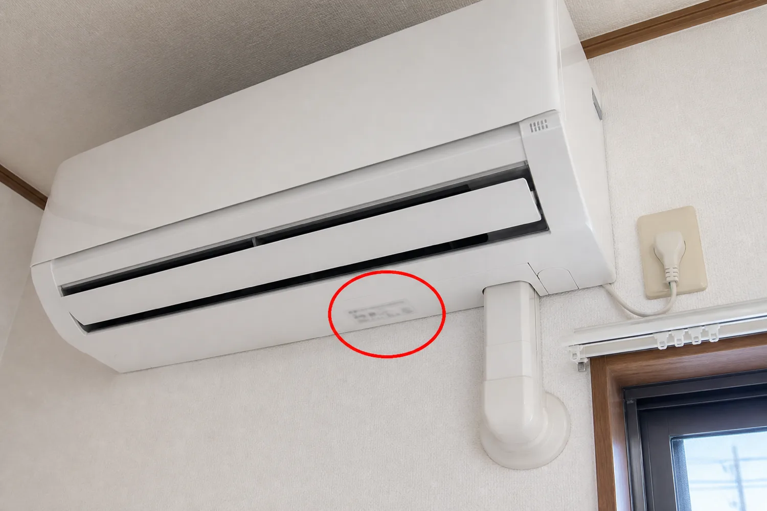 壁掛けエアコンを見上げた角度から、本体下面にある品番シールの位置を赤い丸で示した案内写真