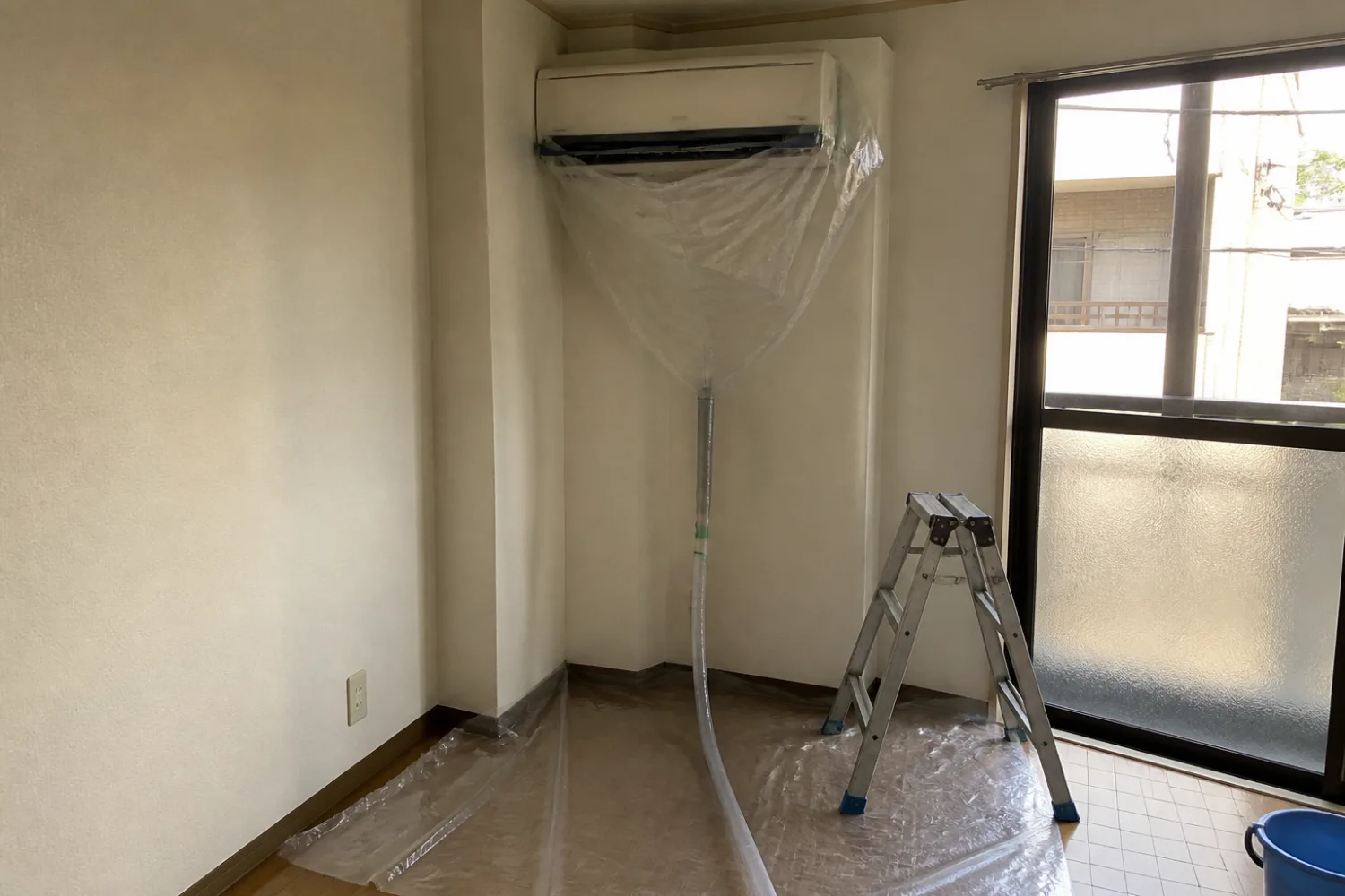壁掛けエアコンに洗浄カバーとホースを取り付け、床を養生したエアコンクリーニングの準備風景