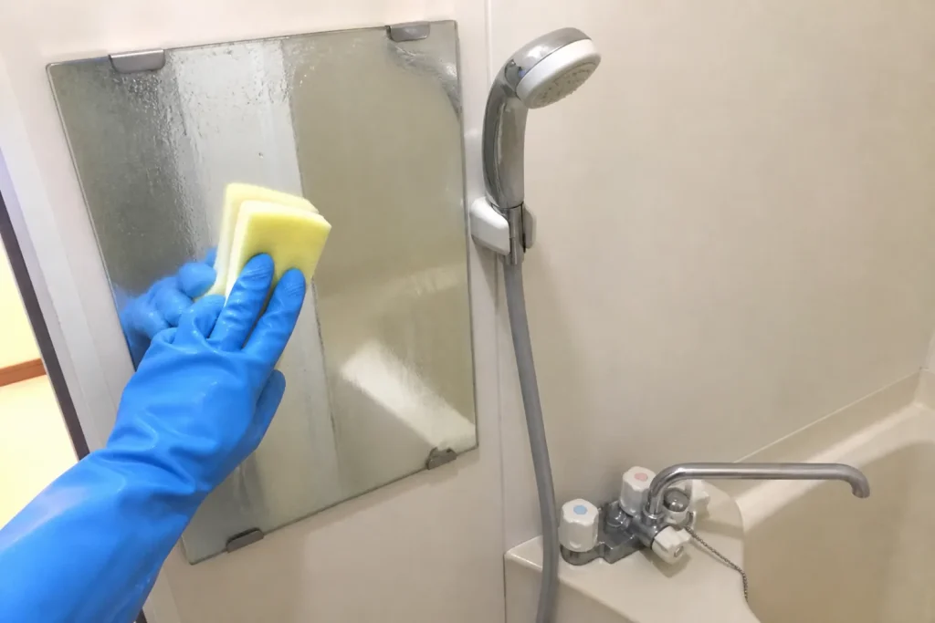 浴室の鏡を青いゴム手袋の手でスポンジ洗いしている。横にシャワーと水栓が見える