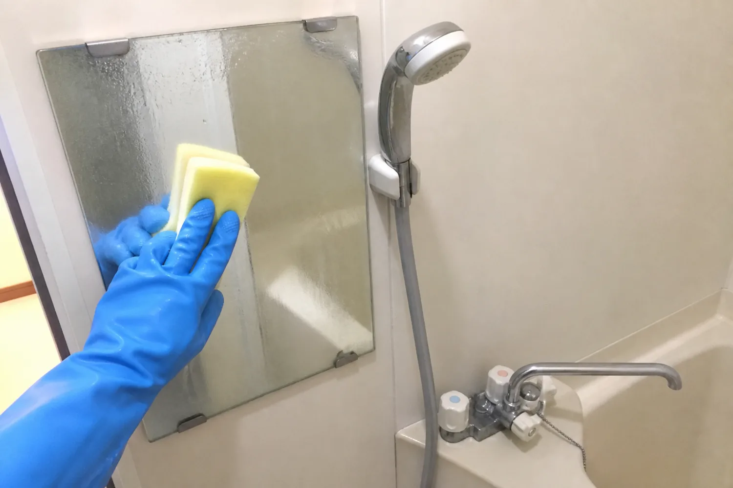 浴室の鏡を青いゴム手袋の手でスポンジ洗いしている。横にシャワーと水栓が見える