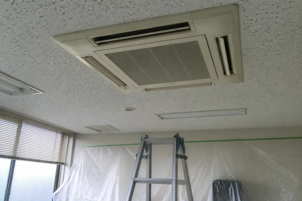 オフィスの天井に埋め込まれた四方向吹き出しの業務用エアコン。下に脚立と養生シートを設置
