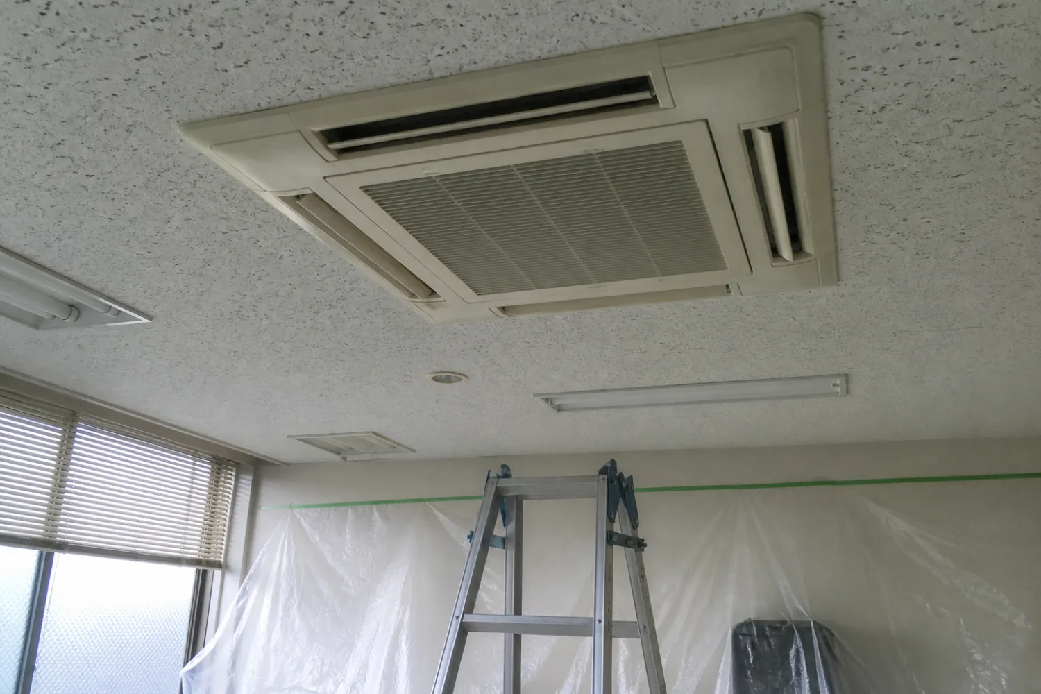 オフィスの天井に埋め込まれた四方向吹き出しの業務用エアコン。下に脚立と養生シートを設置